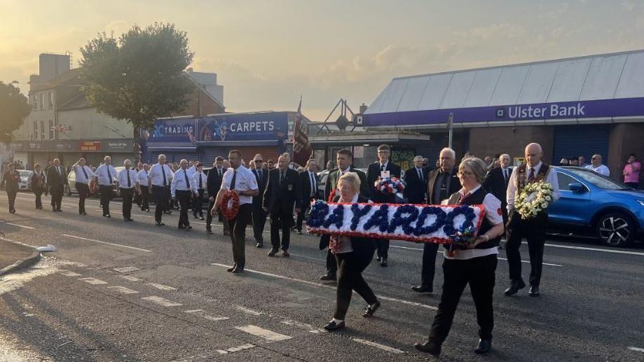 Two women carry a wreath with the words Bayardo on it in blue on a white background. A man is behind them carrying a yellow floral tribute. There are people behind them dressed in black and white.