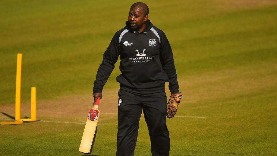 Mark Alleyne, holding a bat in one hand and wearing a glove on the other, while wearing a black Gloucestershire training outfit