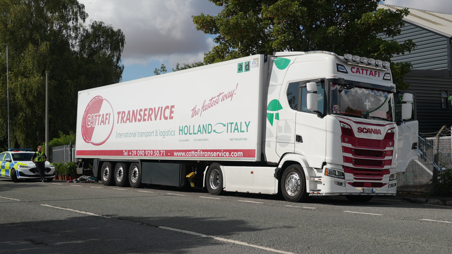 The photo shows a predominantly white articulated lorry, which is stationary. It has red lettering on the side with the name of the haulage company, Cattafi Transervice, and green lettering which says International transport & logistics Holland Italy. Behind the lorry, there is a police car and a police officer. 