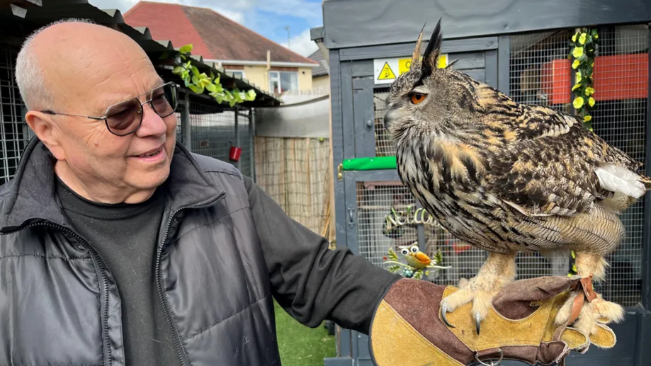 A man in a black jumper and puffer jacket smiles in a garden, as an owl sits on his hand in a bird handling glove