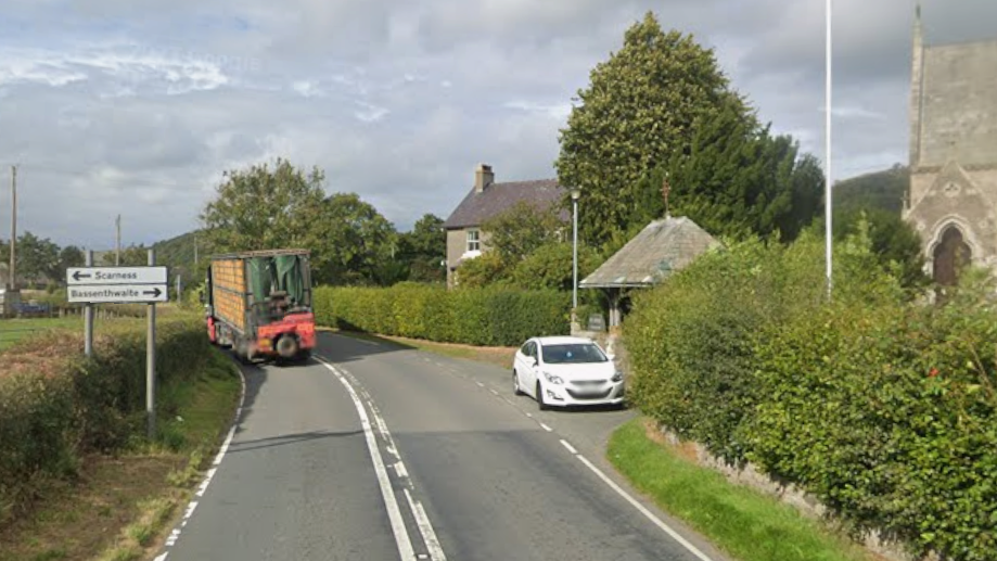 The A591 road is a singe carriageway at Chapel near Bassenthwaite. There is a farm truck travelling one way and a white car parked outside a church on the right. There are green bushes lining the road.