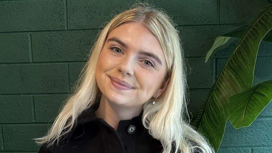 A head and shoulders shot of Aleasha, who is smiling into the camera in front of a painted green wall. She has blonde hair down over her shoulders and is wearing a black jacket unbuttoned at the collar.