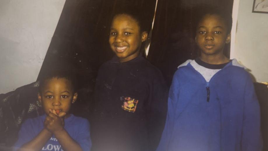 An old photo that shows a very young Joe Aribo (left) as a toddler wearing a blue t-shirt alongside his elder brother and sister. His sister Antonia is in the centre of the photo in a dark blue top smiling into the camera while his brother Paul wears a neutral expression on his face and a baggy blue zip-up top