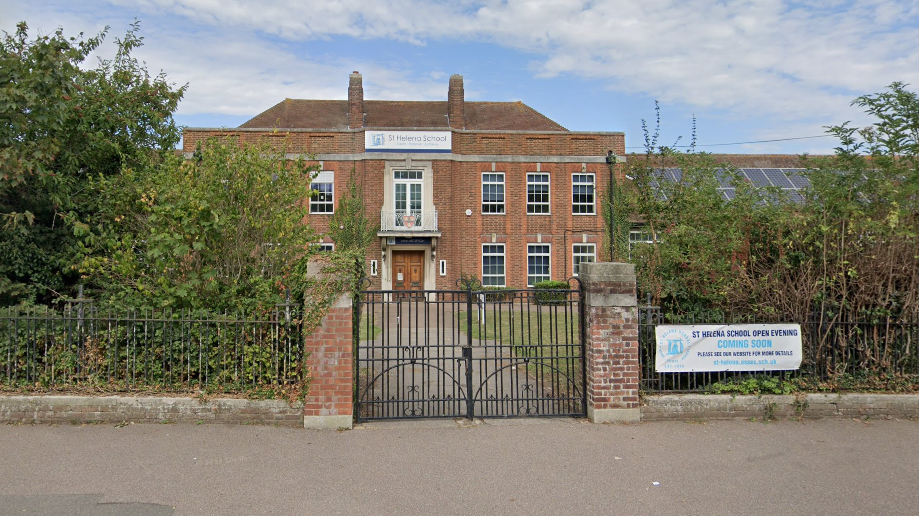 The exterior of the school building. It is mostly brick with large windows and a balcony. Looks early 20th Century. There are black gates with a drive leading to entrance.