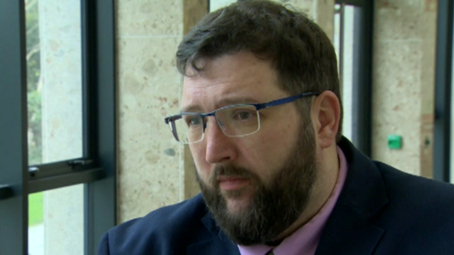 Dr Perry has short dark hair, silver glasses with blue, and dark facial hair, He is wearing a pink shirt with a silver tie and a navy suit jacket. Behind him is a black window and a stone wall.