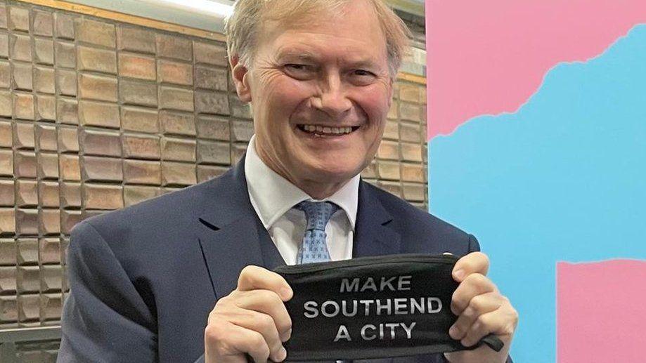 Sir David Amess holding a face mask that has "make Southend a city" printed in white font on it. Sir David is wearing a navy suit and tie and is standing in front of a brick wall.