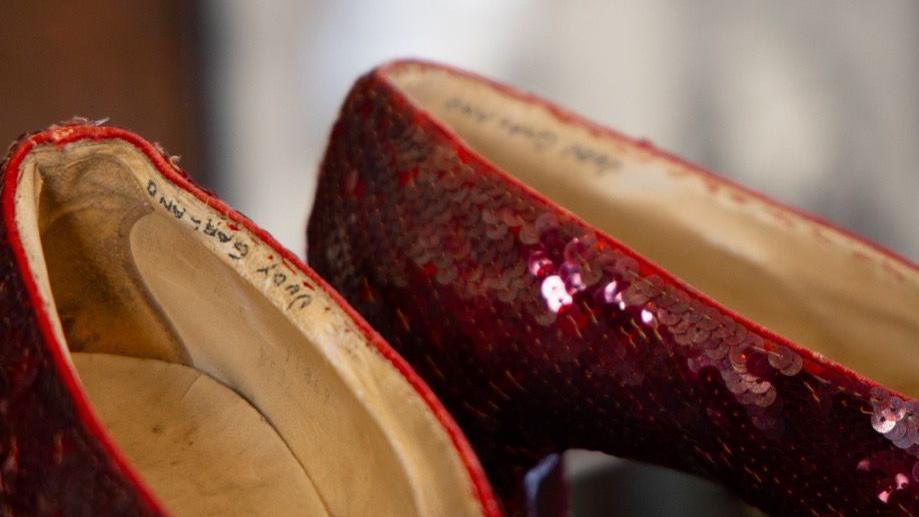 A close-up of the ruby red sequin shoes which feature dark red sequins sewn in diagonal strips across the side of the shoes, with a light coloured insole. The name 'Judy Garland' is written in capitals in black pen on the inside seam of each shoe