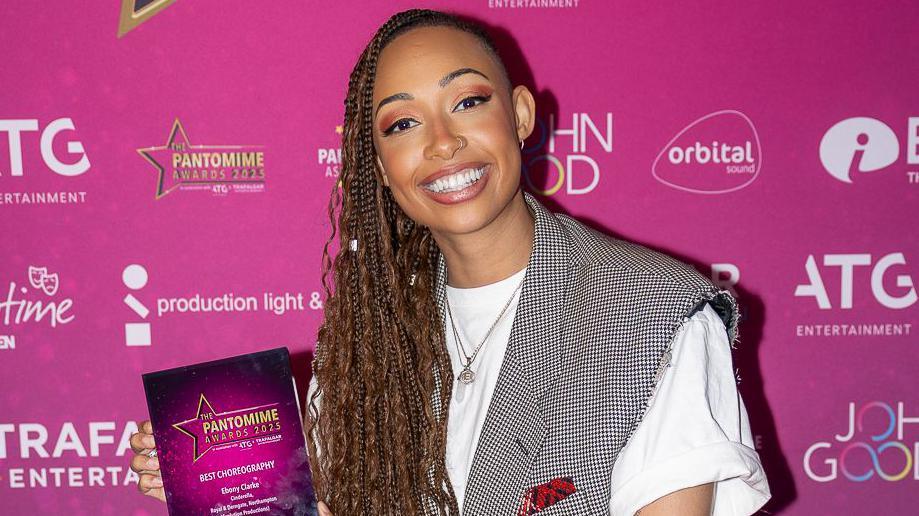 Ebony Clarke holding a pantomime award, in front of a pink back drop. Ebony is smiling, straight at the camera, wearing a white top and checked jacket, she has long dark hair in braids. She has a nose ring, a necklace on and a hoop earring in her left ear.