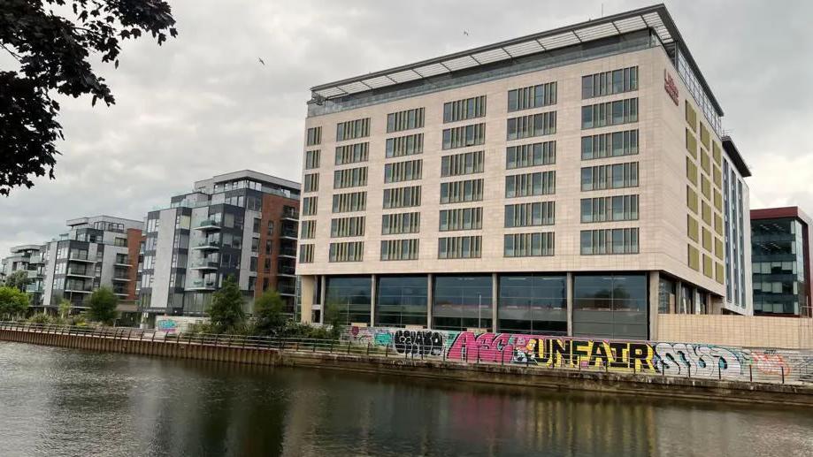 Side profile picture of the Hilton Hotel by the River Nene- a tall building with glass windows next to blocks of apartments.