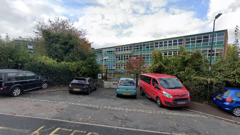A derelict building. The building is a large green structure with lots of windows, many of which are broken. The building is shrouded by trees. Cars are parked in front gates in front of of the building.