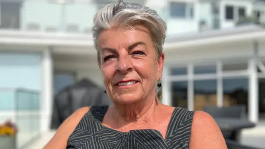 Penny Bromley smiles while sitting in a chair in a garden. She is wearing a black and grey dress and has black dangly ear-rings.