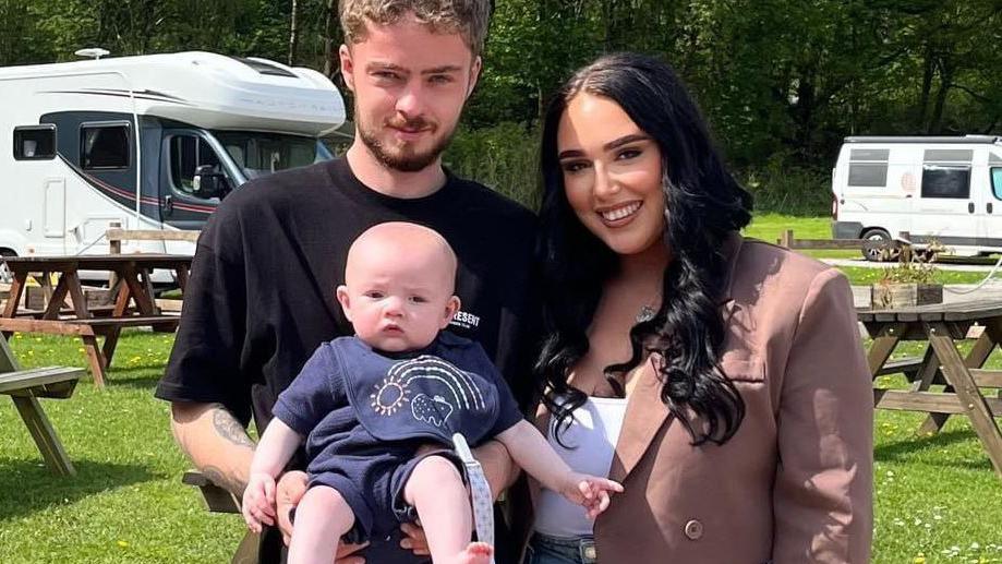 A young man with fair hair and wearing a black t-shirt holds a baby. The baby is wearing a navy outfit. A woman with long dark hair and wearing a brown blazer and white top stands next to them. In the background on the grass are some picnic benches and two caravans.