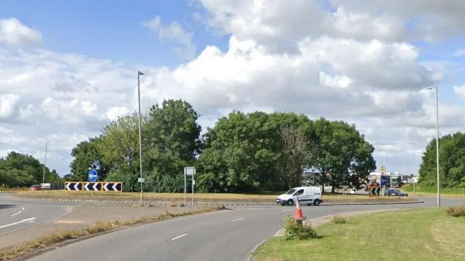 A roundabout with trees in the middle. A white van is being driven around it. 