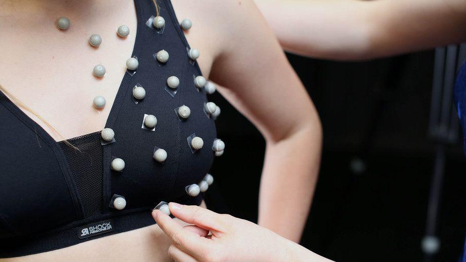 A close up of a woman's chest wearing a dark coloured sports bra with chickpea-sized grey spheres being stuck on in a rows and columns across the bra.
