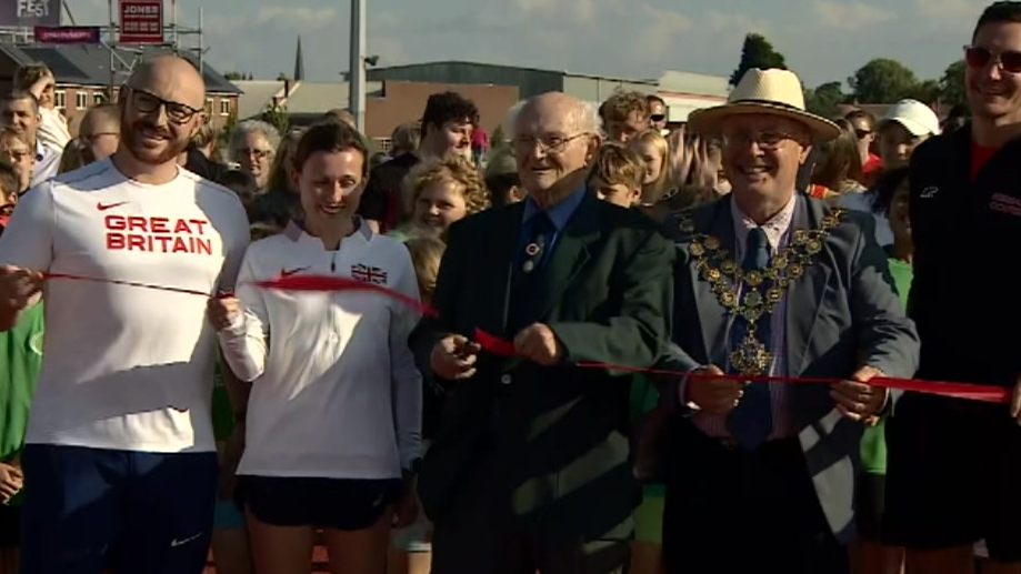 Three men and a woman stand on the track cutting a red ribbon, with crowds of people behind them