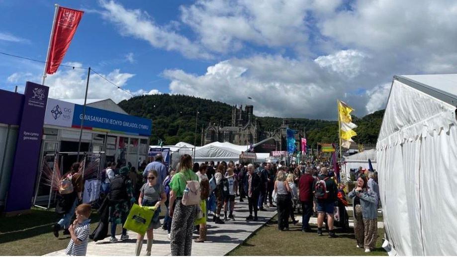 Crowds of people at this year's Eisteddfod. There are stalls on either side of the path in white tents, with Margam Castle behind.