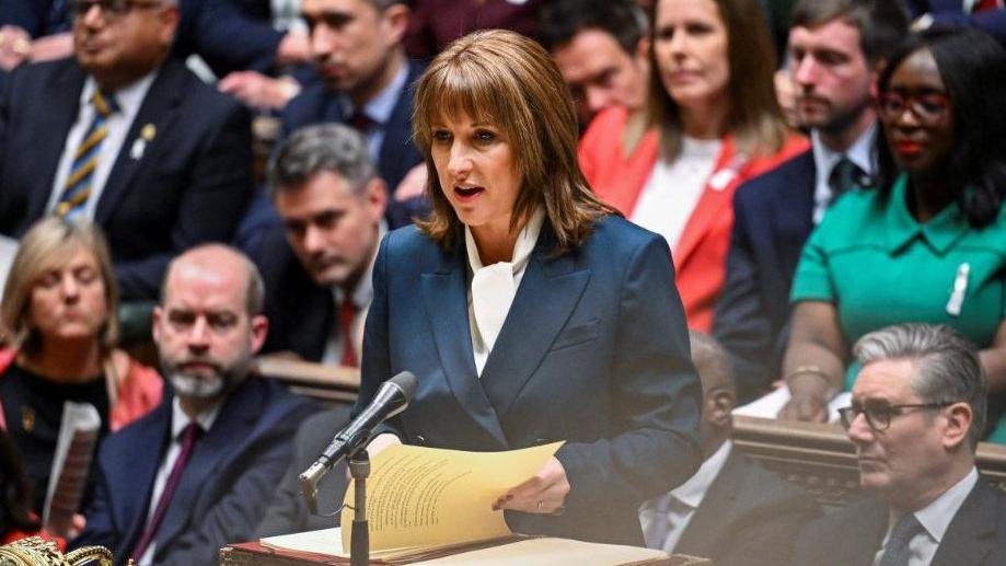 Rachel Reeves announces the Budget standing at the dispatch box in the House of Commons. She has shoulder-length brown hair with a fringe and is wearing a navy suit with a white shirt. Dozens of Labour MPs are sitting behind her.
