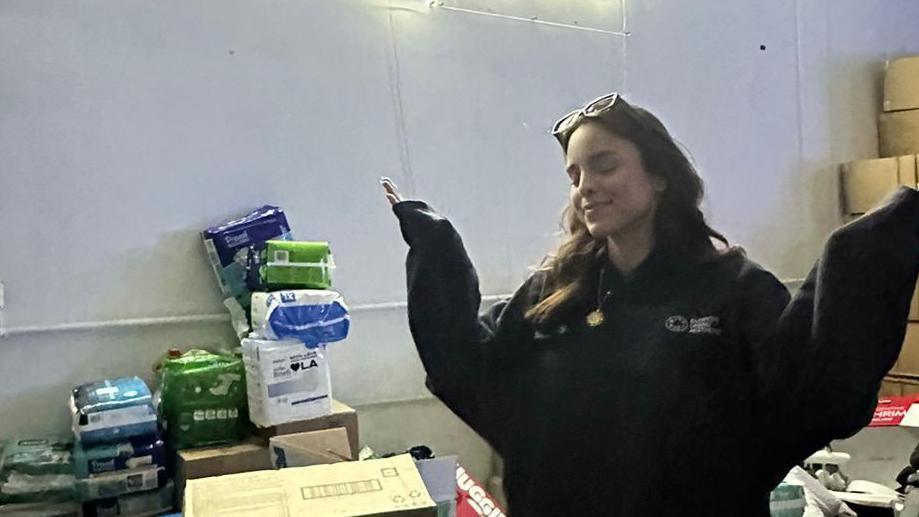 Charlotte Hawthorne poses in an LA evacuation centre. She has long dark hair with glasses that rest on the top of her head. She also wears a dark coloured hoodie and has her arms raised.