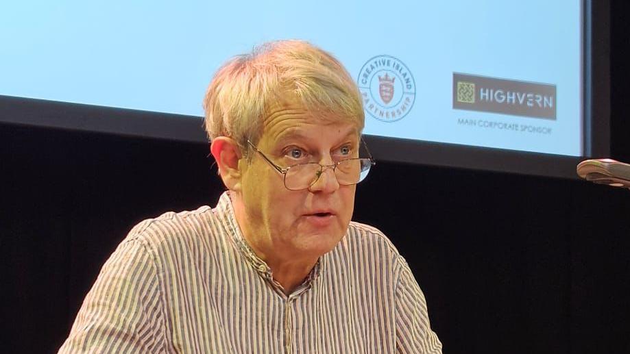 A man, in a white and purple striped top. He is looking forwards and his mouth is slightly open. He is wearing glasses. Behind him is a projector screen and black curtain. 