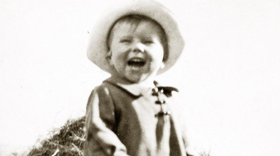 A small boy in a brimmed hat and an old-fashioned coat with a collar wears an expression of joy, with his mouth wide open