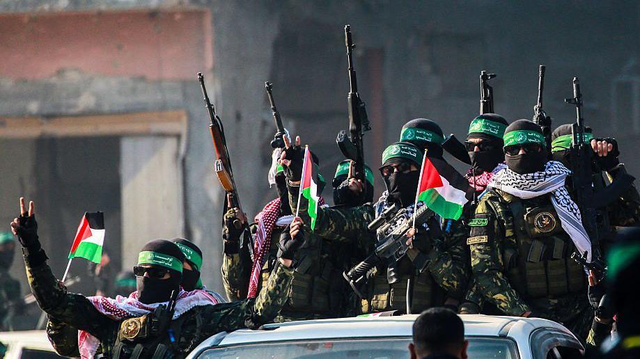 A number of armed Hamas militants arrive in an area before releasing Israeli hostage Agam Berger to a Red Cross team in Jabalia, Gaza on 30 January 2025, as part of a hostage-prisoner exchange. The fighters wear black sunglasses, black balaclavas, green headbands and keffiyeh scarves. Most of them carry guns and three are also waving Palestinian flags. 