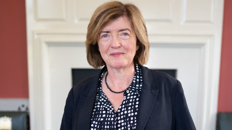 Sue Gray wearing a navy blouse with white spots, a small, beaded navy necklace, and a navy jacket smiles slightly in a Downing Street office.