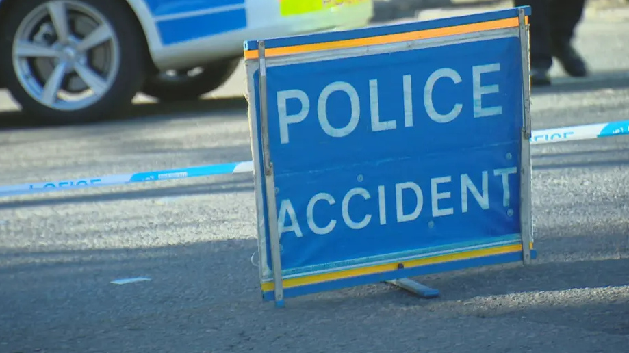 A blue sign in the middle of the road. It has the words 'Police Accident' in white letters. The sign is in front of blue and white police tape and the wheel of a police car is visible in the background.