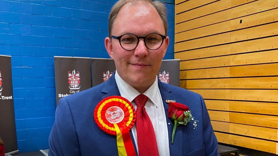 A headshot of a man wearing a blue suit, with white shirt and red tie. He is smiling into the camera and standing in a sports hall. He has circular, bold glasses and is wearing a red rose and red rosette on his blue suit. The rosette reads his name, Gareth Snell. 