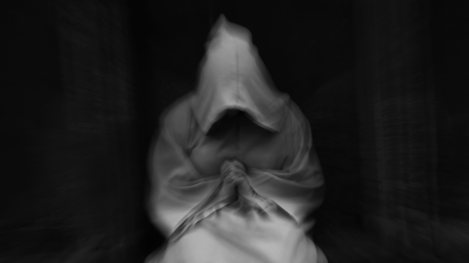 A blurry black and white photo of a hooded figure clasping its hands in a dark room.