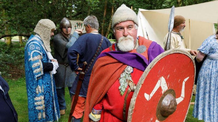 A man dress as a Viking holds a red shield with with a triskelion. Others in traditional dress, including a man in a wig, stand around behind him.