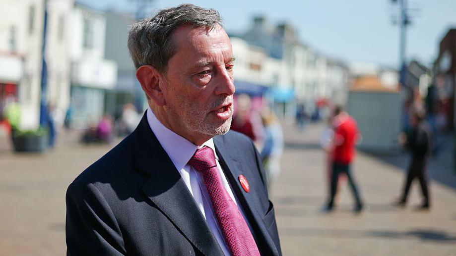 A man in a black suit with white shirt and pink tie stands on a seafront promenade in front of white shops and houses.