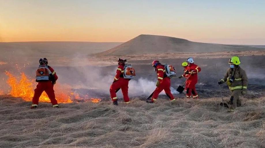 Six firefighters in red overalls tackle a fire on a grassy area of moorland 