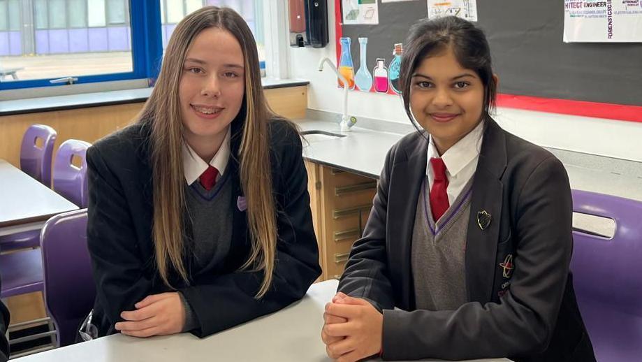 Emilia and Dewmi are sitting on purple seats at a white table in a classroom. Emilia is smiling with long brown hair and is wearing her school uniform with a red tie, a grey jumper and black blazer. Demwi is also smiling and her black hair is tied back in a bun. 