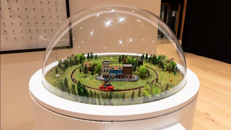 A mini model village is surrounded by trees and grass and a cartoon character is riding a scooter around a narrow road encompassing the village. The exhibit is covered in a glass dome.