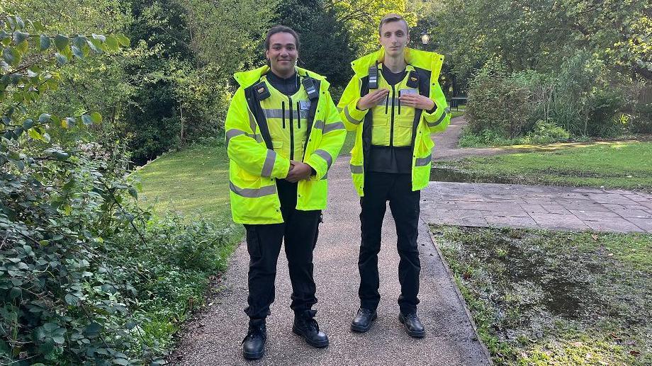 Street wardens Jayden Jermy and Dalton Campen stand side by side in a park. They wear high visibility jackets black trousers and big boots. Jayden has shoulder length dreadlocks tied neatly in a ponytail. Dalton is taller and has blonde hair. 