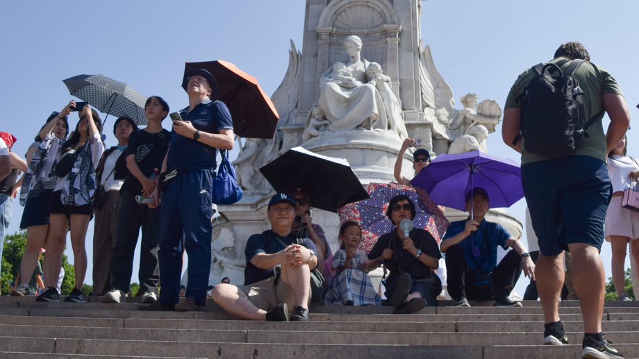 A group of people shield from the sun under umbrellas next to the Victoria Memorial outside Buckingham Palace