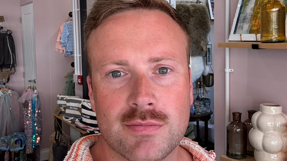 Rowan Hunter standing in front of shelves of bags, clothes and pots inside his shop. He has a moustache and stubble, as well as short fair hair. He is smiling.