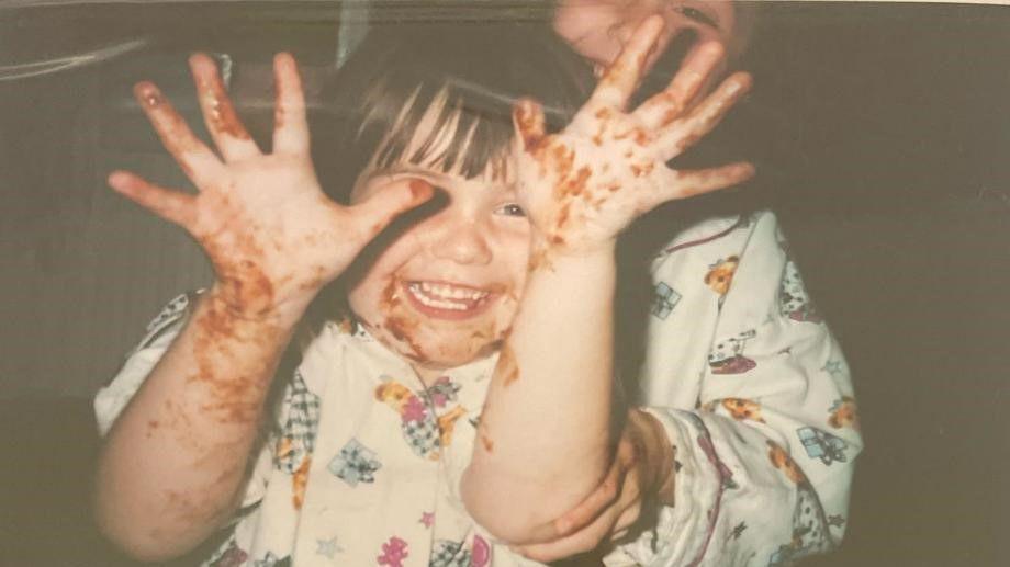 An old-style photo of Courtney Mitchell as a toddler. She has what appears to be chocolate on her hands and she is smiling.