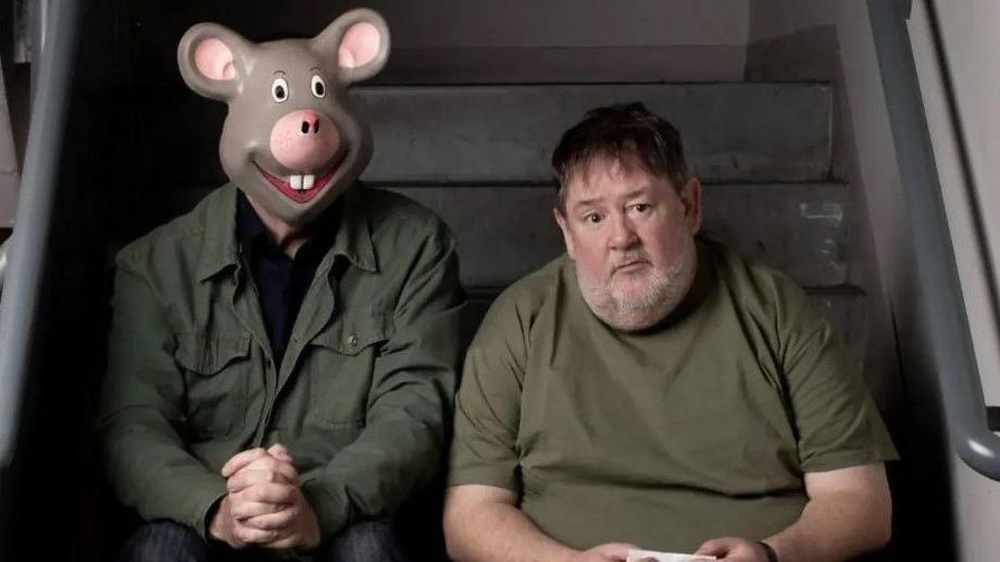 Johnny Vegas sits glumly next to a tall man wearing a mask of a cartoon mouse's head over his face. Both are wearing dark green tops and are sitting on the steps of a staircase.