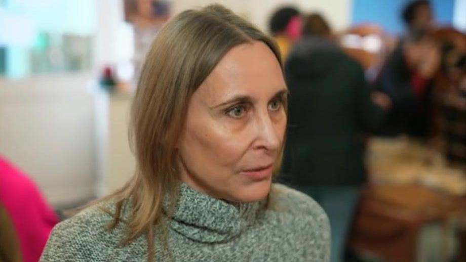 A woman with shoulder-length blond hair wearing a grey knitted turtle neck looking past the camera.