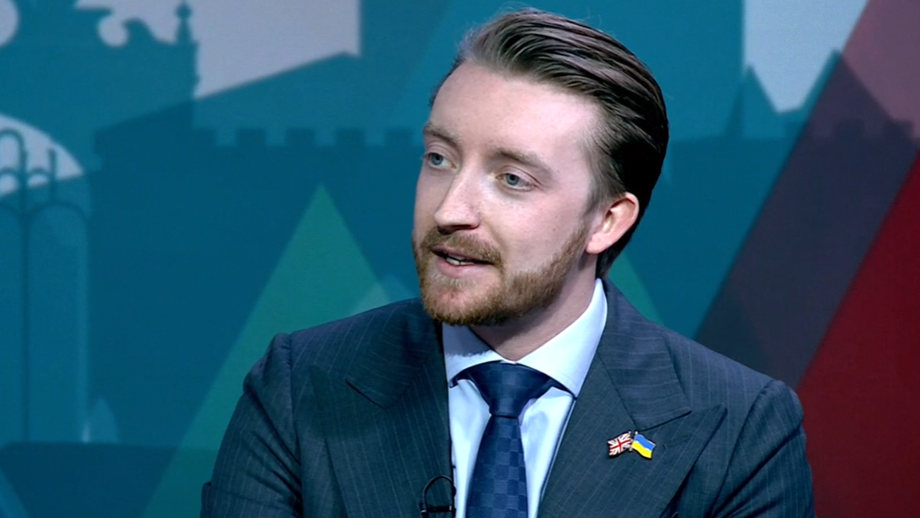 Jaymey McIvor is pictured from the shoulders upwards. He is wearing a blue suit jacket, a blue shirt, and blue tie. Has has two pins on the left side of the jacket, one is a union jack, and the other is a flag of Ukraine. He has brown hair and beard.