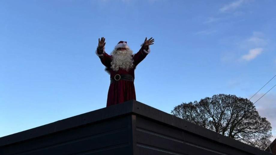 Santa dressed in a red coat, a black belt and a red and white hat with a white beard stood on the roof of the school. The sky is blue.