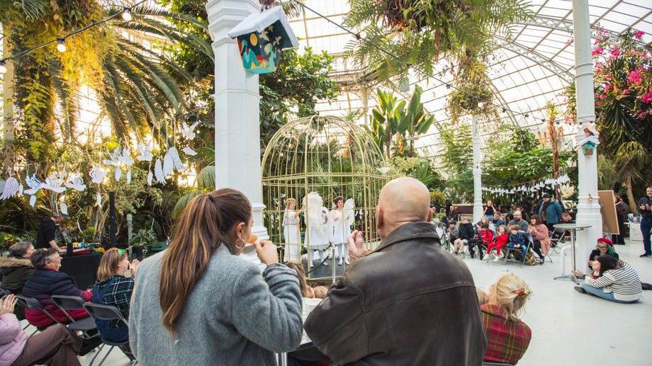 People pictured sitting and standing in the Palm House as they watch a performance inside the Victorian Palm House. In the centre, there are people dressed as angels performing in a gold cage. 