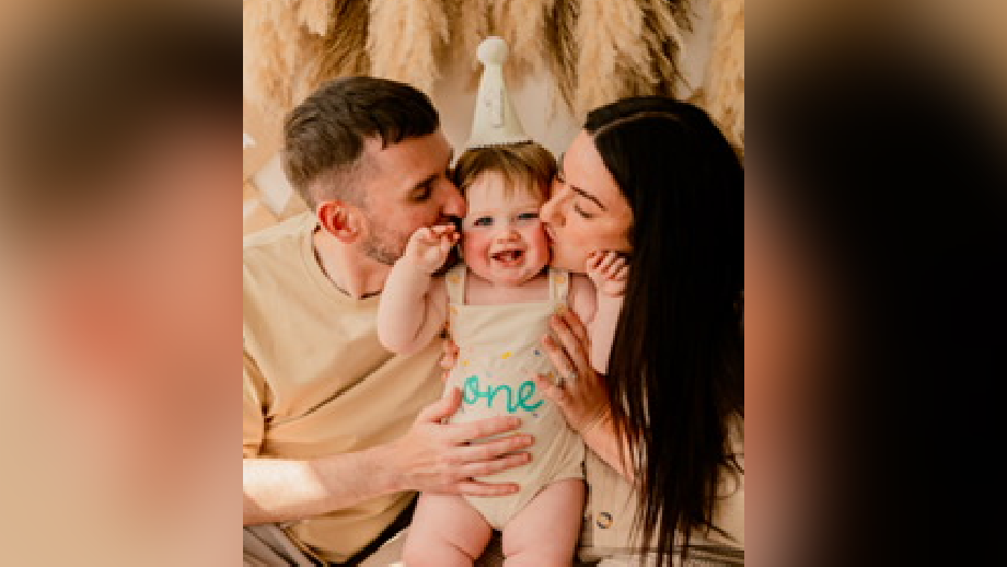 Family picture of Claire and her husband holding Osian at his first birthday party. Osian is in the middle and is wearing a beige birthday hat and a beige baby grow with the word 'one' in green letters on. He is smiling. Claire's husband is on the left and is kissing Osian. He is wearing a beige t-shirt. Claire, on the right, is also wearing a beige blouse.