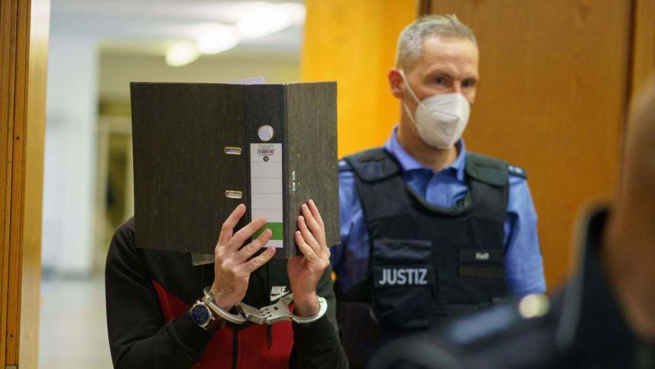 Taha al-Jumailly (L) holds a folder in front of his face as he is led into the courtroom for the sentencing in his trial for charges of genocide, crimes against humanity, war crimes, human trafficking and murder at the Higher Regional Court in Frankfurt am Main, Germany (30 November 2021)