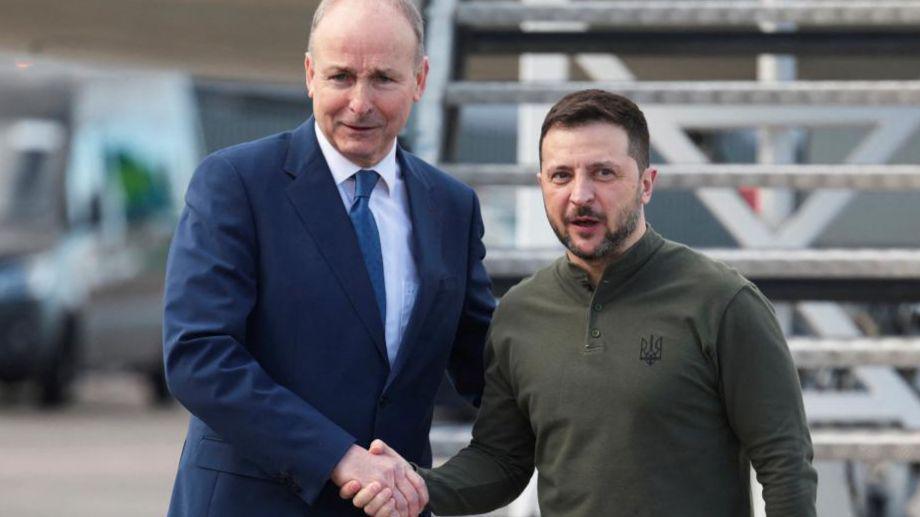 Volodymyr Zelensky and Micheál Martin shake hands at the steps of a plane at Shannon Airport. Martin is wearing a navy suit with a white shirt and blue tie. Zelensky has a black and grey beard and is wearing a green long sleeve top with a Ukrainian symbol.