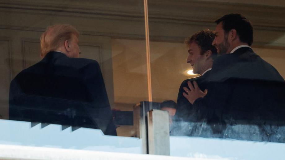 Donald Trump, Daniel Penny and JD Vance speak in a suite while attending an Army v Navy American football game in Landover, Maryland. All three have their backs to the camera, with their faces only partially visible. Vance has his hand on Mr Penny's shoulder, and both appear to be conversing with Trump.