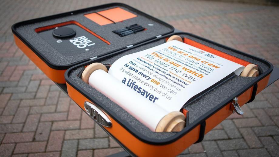 An orange box containing a scroll. The scroll is white with blue and orange writing on. Next to it is speaker. The pledge reads: Whoever we are, wherever we are from, we are one crew, ready to save lives. We’re powered by passion, talent and kindness, like generations of selfless lifesavers before us. This is our watch, we lead the way, valuing each other, trusting each other, depending on one another, volunteering to face the storm together. Knowing that, with courage, nothing is impossible. That is what has always driven us to save every one we can. It's what makes every one of us a lifesaver.