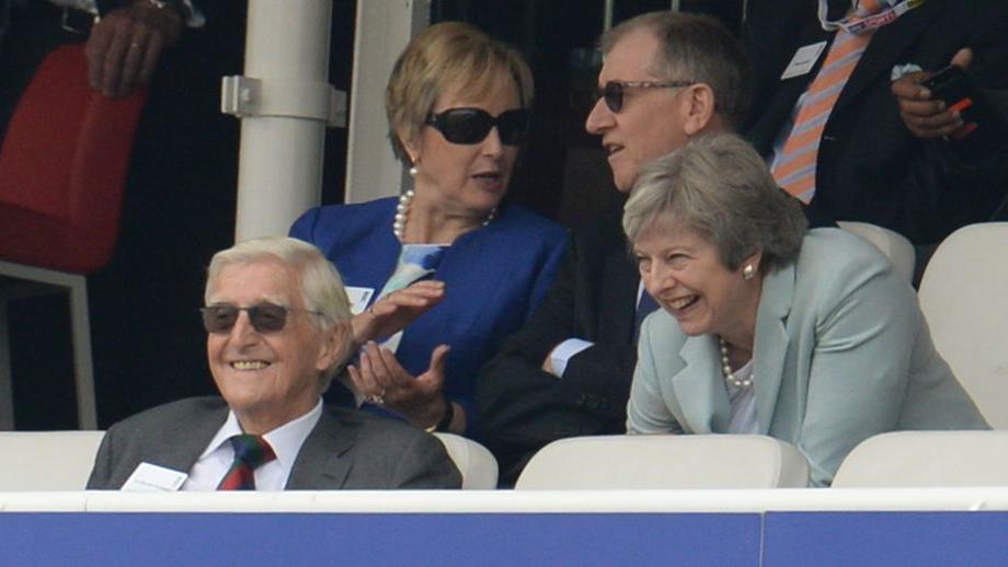 Sir Michael Parkinson sat in the front row of a seated area at Lord's, with Theresa May sat behind him, them both laughing. Mrs May's husband Philip is sat to her right 