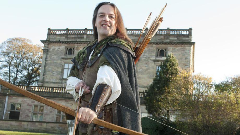 A man with long hair and green robes holding a bow and arrow. He stands in front of Nottingham Castle